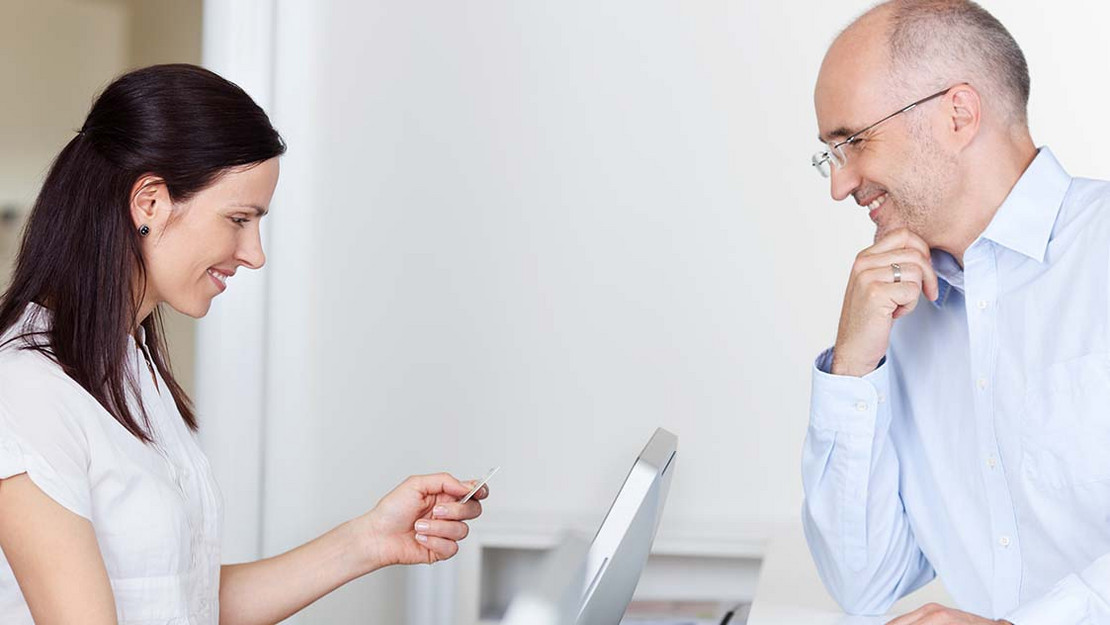 Frau am Schalter einer Praxis hält die Versichertenkarte eines Patienten in der Hand. 
