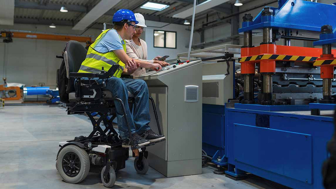 Mann in einem elektrischen Rollstuhl und eine Frau in einer Fabrik am Bedienpult einer Maschine.
