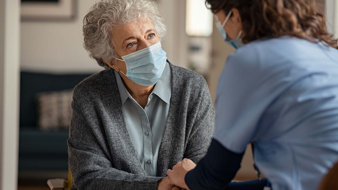 Alte Frau mit Maske und Pflegekraft sitzen sich gegenüber.