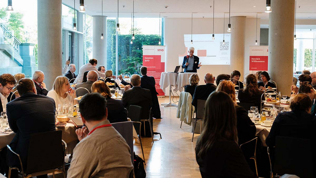 Voller Tagungsraum im Bundestag. Menschen sitzen in Gruppen an Tischen beim Frühstück und verfolgen einen Vortrag mit Präsentation, den Rolf Schmachtenberg hält.