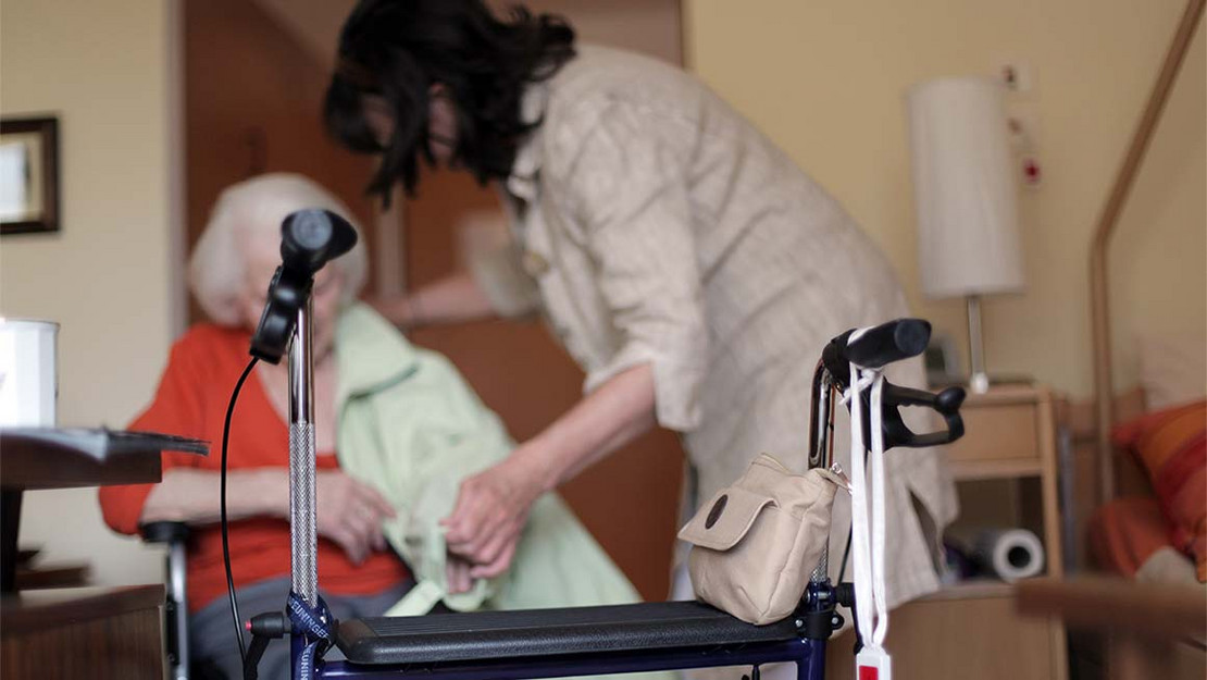 Eine ältere Frau wird von einer jungen Frau im Pflegeheim besucht. Im Vordergrund steht ein Rollator. 