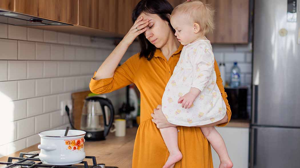 Gestresste Frau mit Kind auf dem Arm kocht am Herd.