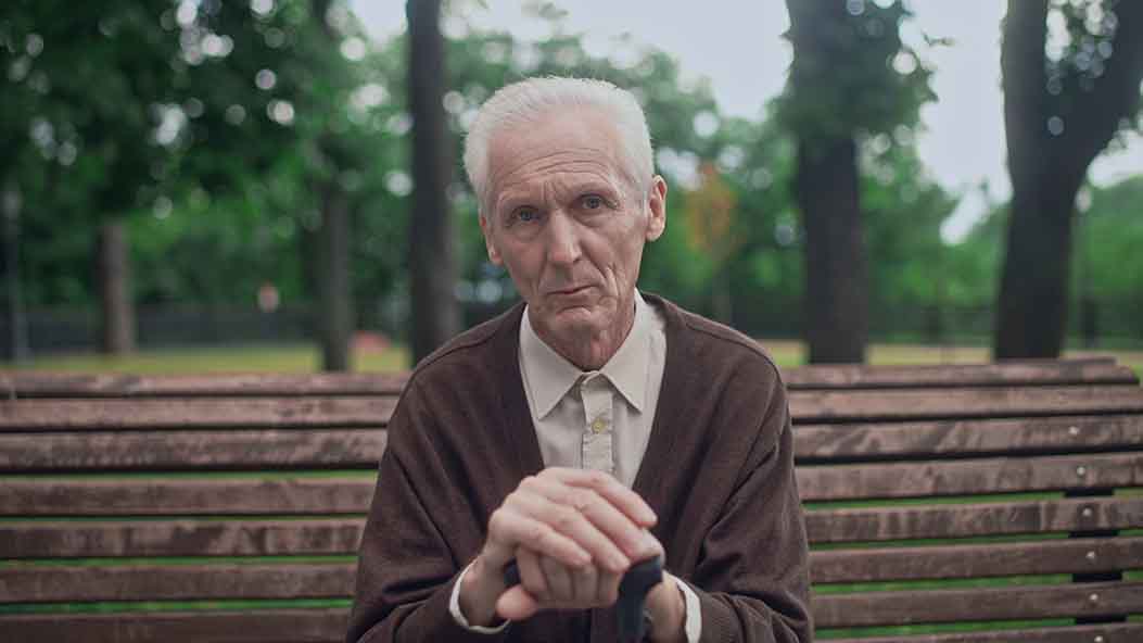 Alter Mann sitzt auf einer Bank und blickt ernst in die Kamera.