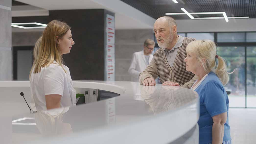 Ein älteres Ehepaar steht an der Rezeption in einem Krankenhaus und spricht mit einer Beschäftigten.